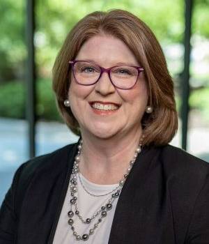female headshot wearing a dark jacket and light tan shirt with necklaces and glasses